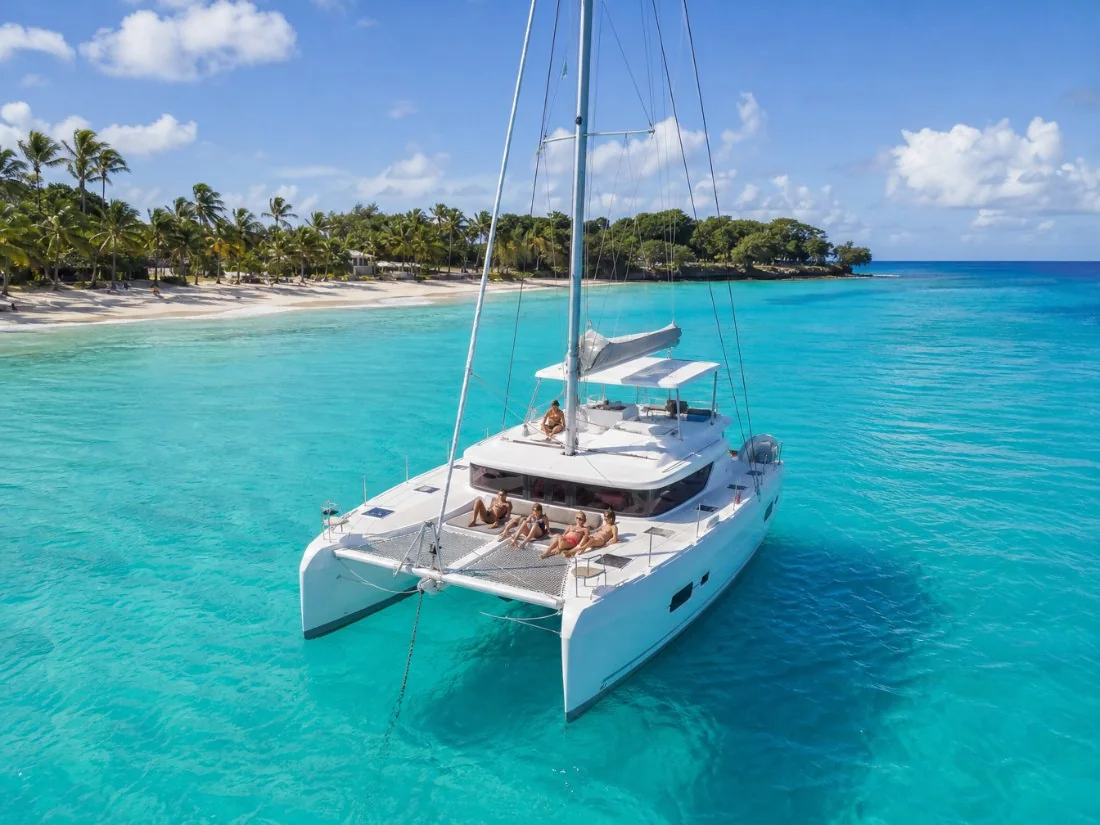 Ein weißer Segelkatamaran mit Gästen auf den Trampolin-Netzen, vor Anker in türkisfarbenem Wasser vor einem palmengesäumten Strand in Barbados